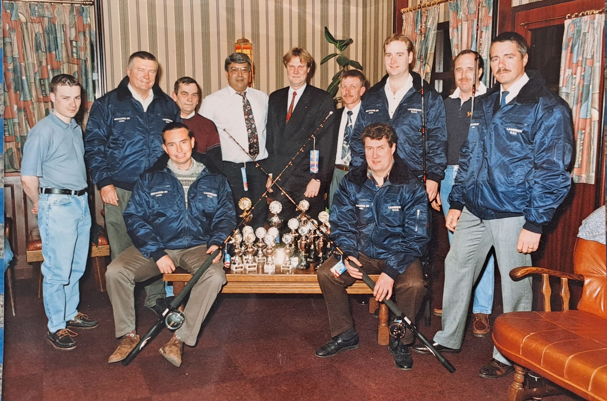 Group of people in blue jackets posing with trophies in a room with bookshelves and a leather chair.